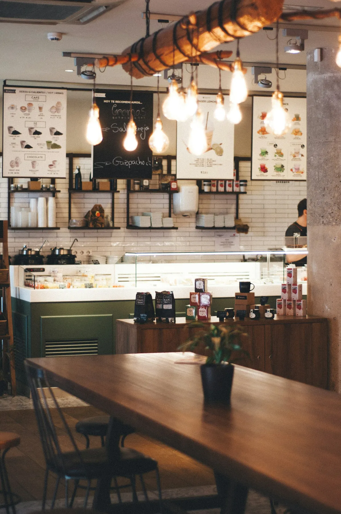 Inside a cosy café with a long wooden table, pendant lights and a counter of bakes