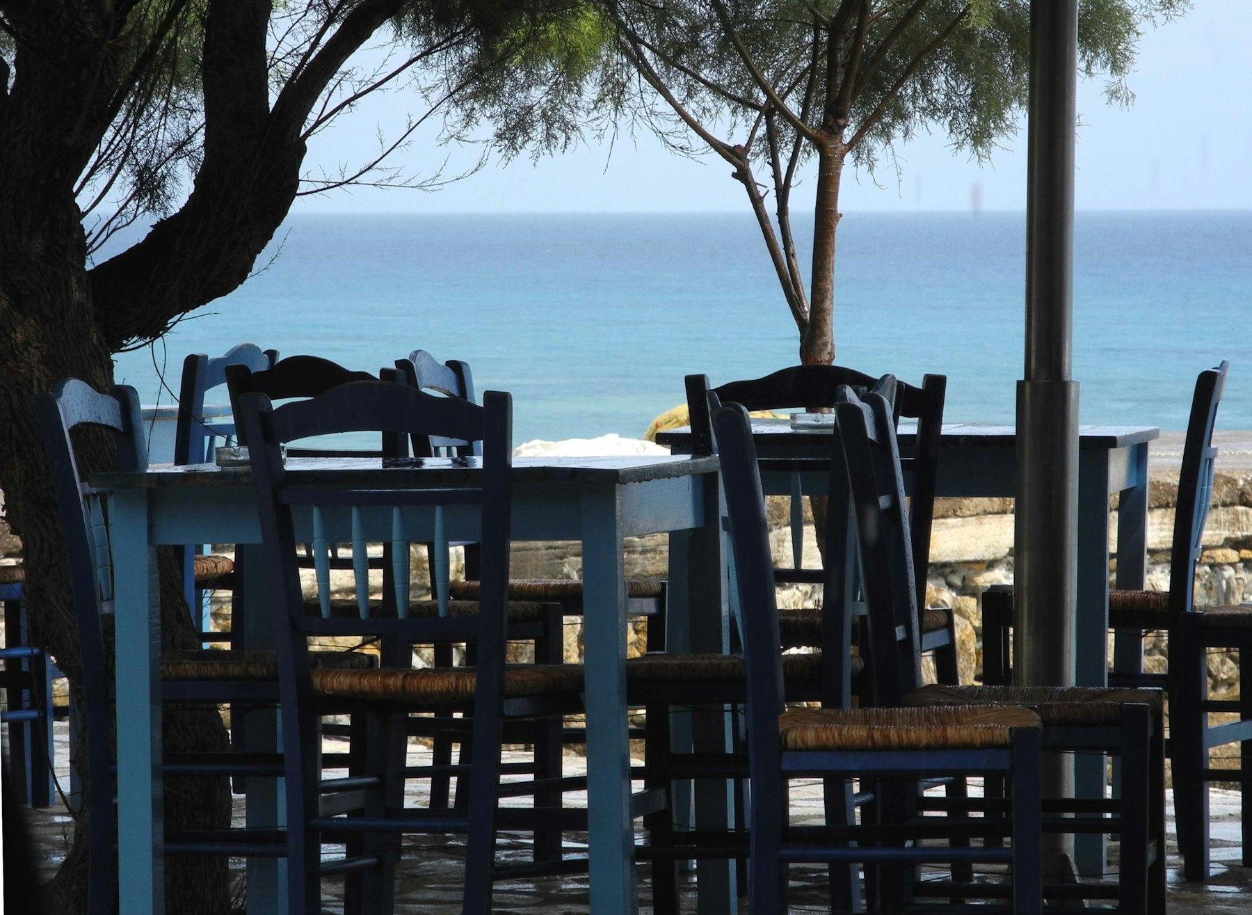 Wooden taverna tables and chairs under a tree, looking out to a blue sea